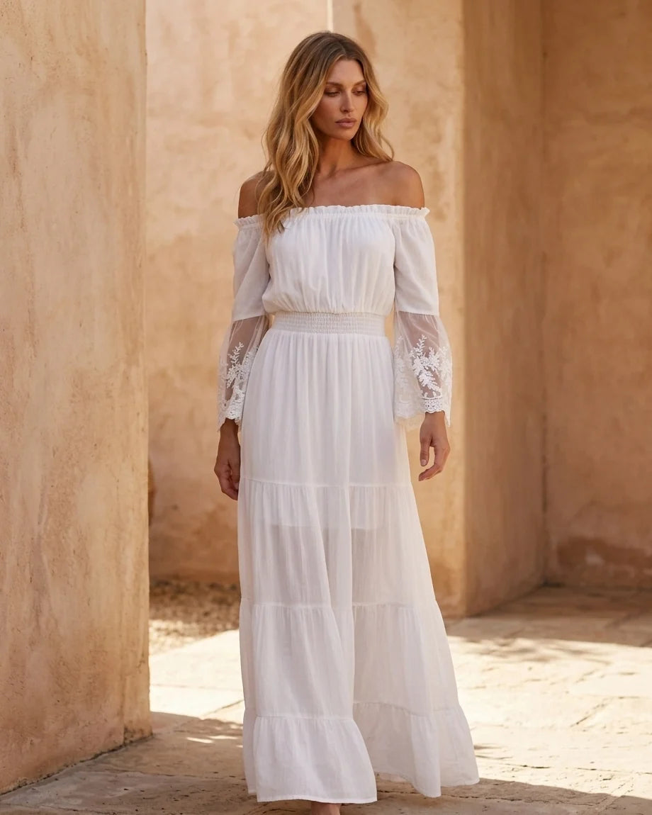 A flowing off-the-shoulder maxi dress in white with lace-trimmed bell sleeves and a tiered skirt.